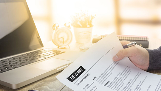 A person holding a resume while sitting at a desk with a laptop and a clock in the background.