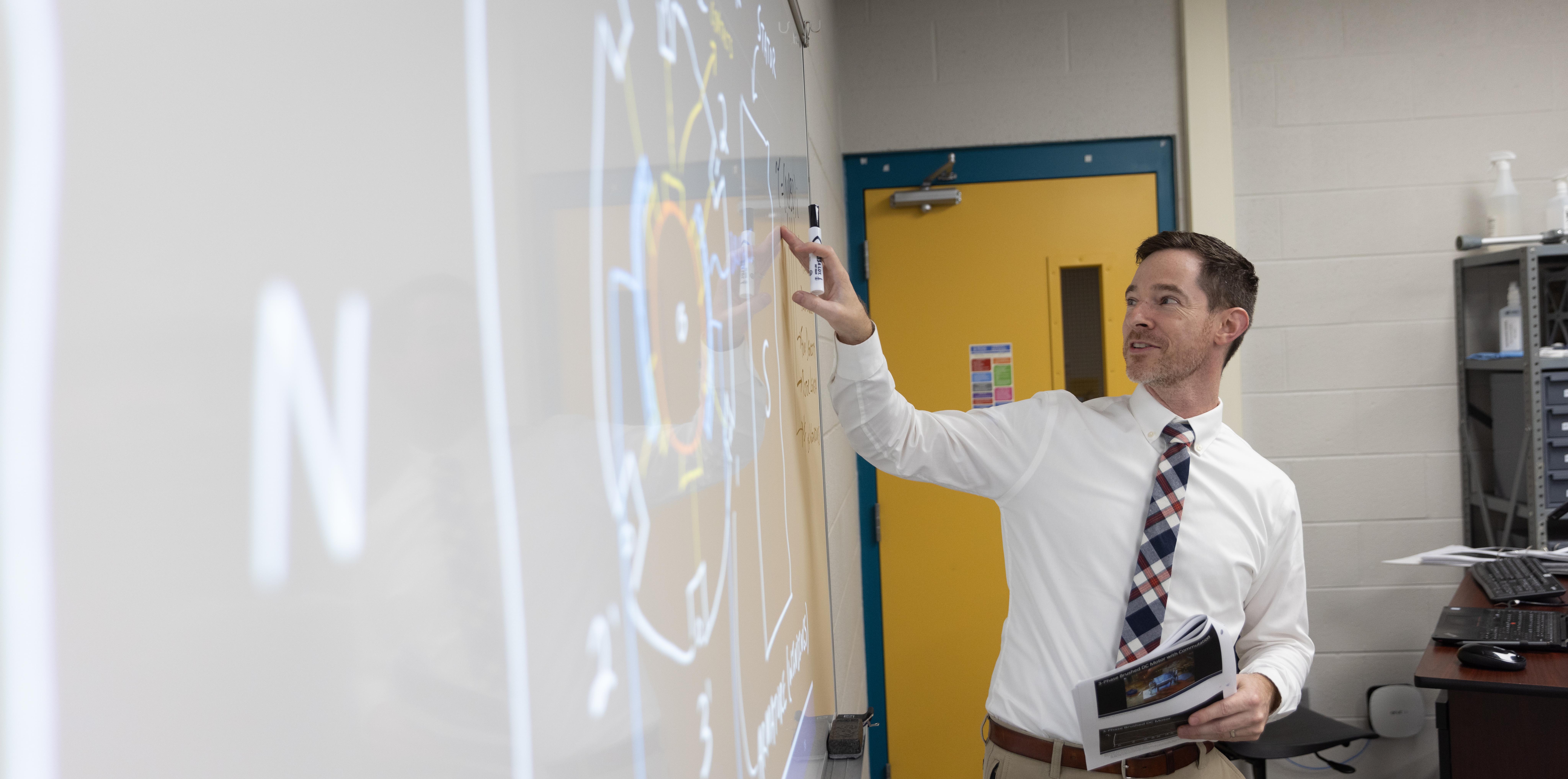 A male teacher stands at a white board, instructing the class.