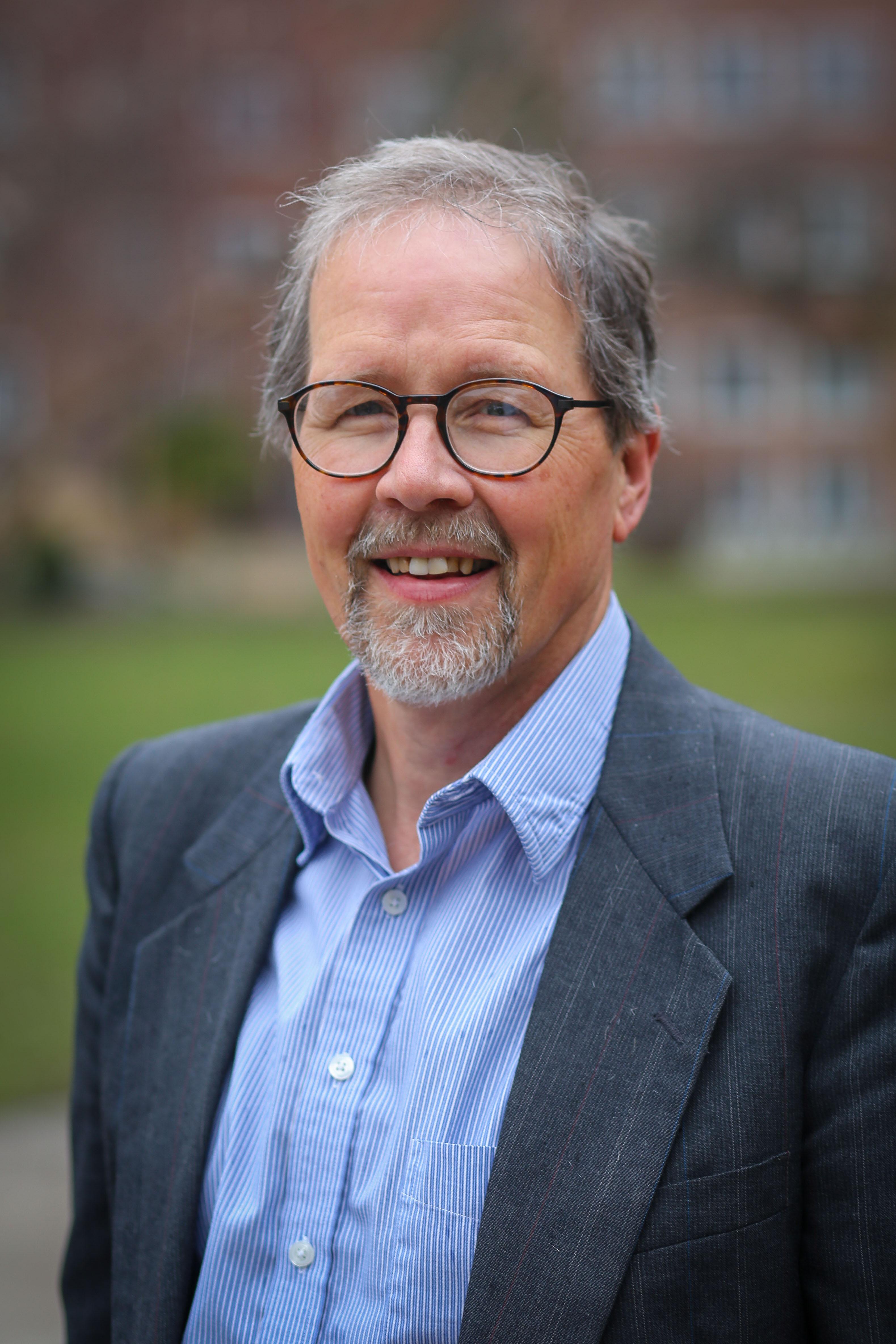 A white man with salt and pepper hair, a goatee, and glasses, smiles at the camera. He is wearing a light blue shirt and a navy blazer.