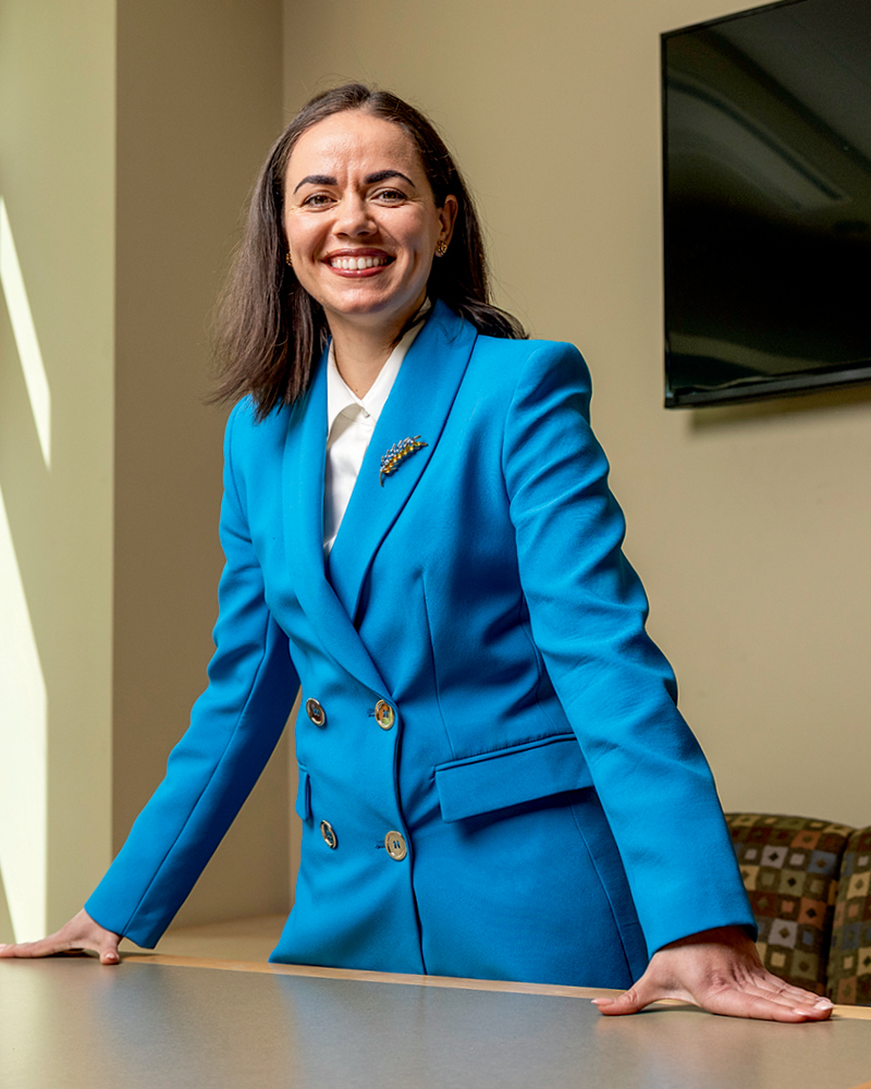 A smiling woman in a bright blue double-breasted blazer stands confidently at a table, exuding professionalism and approachability.