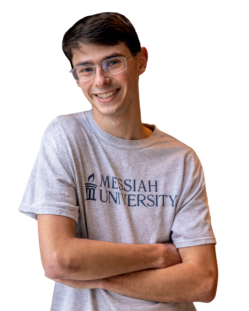 A smiling student wearing a gray Messiah University t-shirt, standing with arms crossed.
