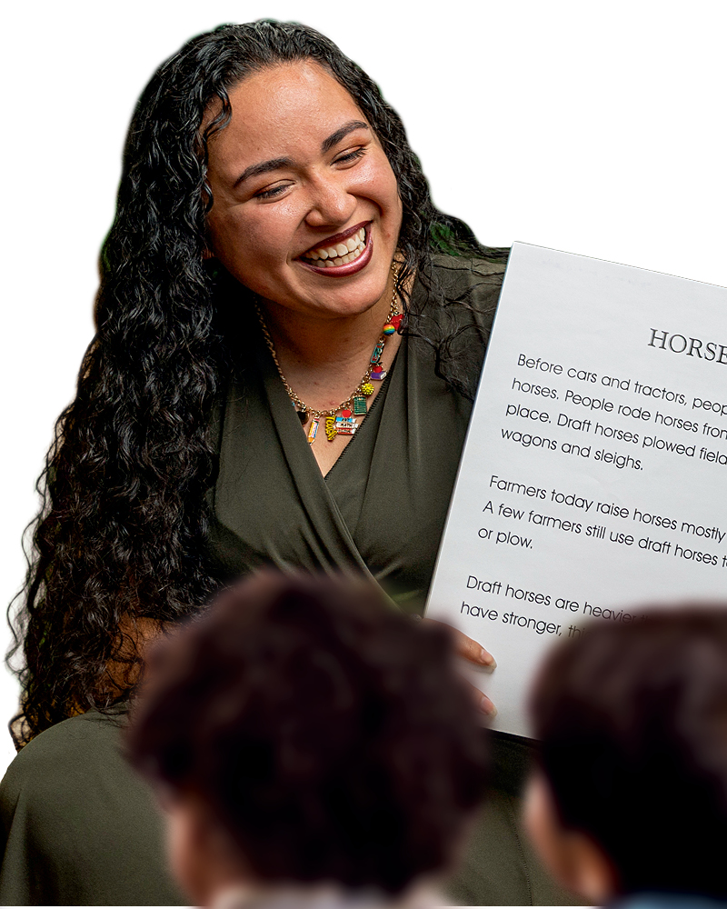 A woman with long dark hair smiles while holding a book, engaging with an audience seen from behind.